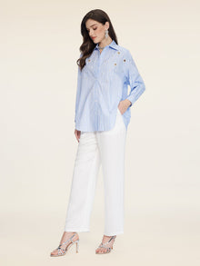 A woman stands against a plain background wearing Social Queen's Pearl & Beaded Striped Blouse with a relaxed fit, paired with white pants and silver heeled sandals.