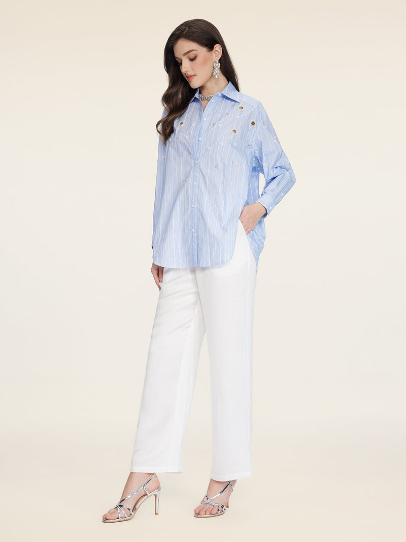 A woman stands against a plain background wearing Social Queen's Pearl & Beaded Striped Blouse with a relaxed fit, paired with white pants and silver heeled sandals.