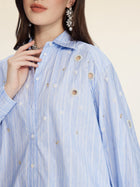 A woman wears the Pearl & Beaded Striped Blouse by Social Queen, featuring light blue stripes, relaxed fit, embellished holes, and hand-applied pearls, styled with statement earrings.