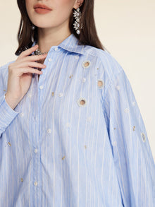 A woman wears the Pearl & Beaded Striped Blouse by Social Queen, featuring light blue stripes, relaxed fit, embellished holes, and hand-applied pearls, styled with statement earrings.