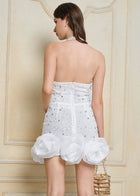 A woman in the Social Queen Pearl-Crystal Embellished Floral Hem Mini Dress faces a textured beige wall, holding a gold purse.