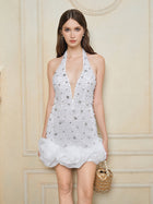 A woman stands before a beige paneled wall in the Social Queen Pearl-Crystal Embellished Floral Hem Mini Dress, featuring a sleeveless white halter silhouette with sparkling details, paired with a gold handbag.