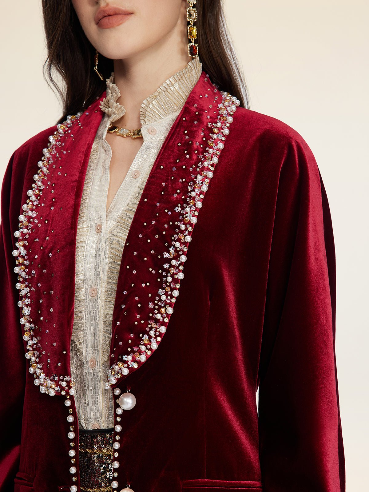 A woman wears Social Queen's Pearl & Crystal Velvet Blazer in deep red, its jeweled trim adding vintage glamour over a textured cream blouse, finished with gold jewelry and drop earrings.
