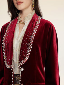 A woman wears Social Queen's Pearl & Crystal Velvet Blazer in deep red, its jeweled trim adding vintage glamour over a textured cream blouse, finished with gold jewelry and drop earrings.