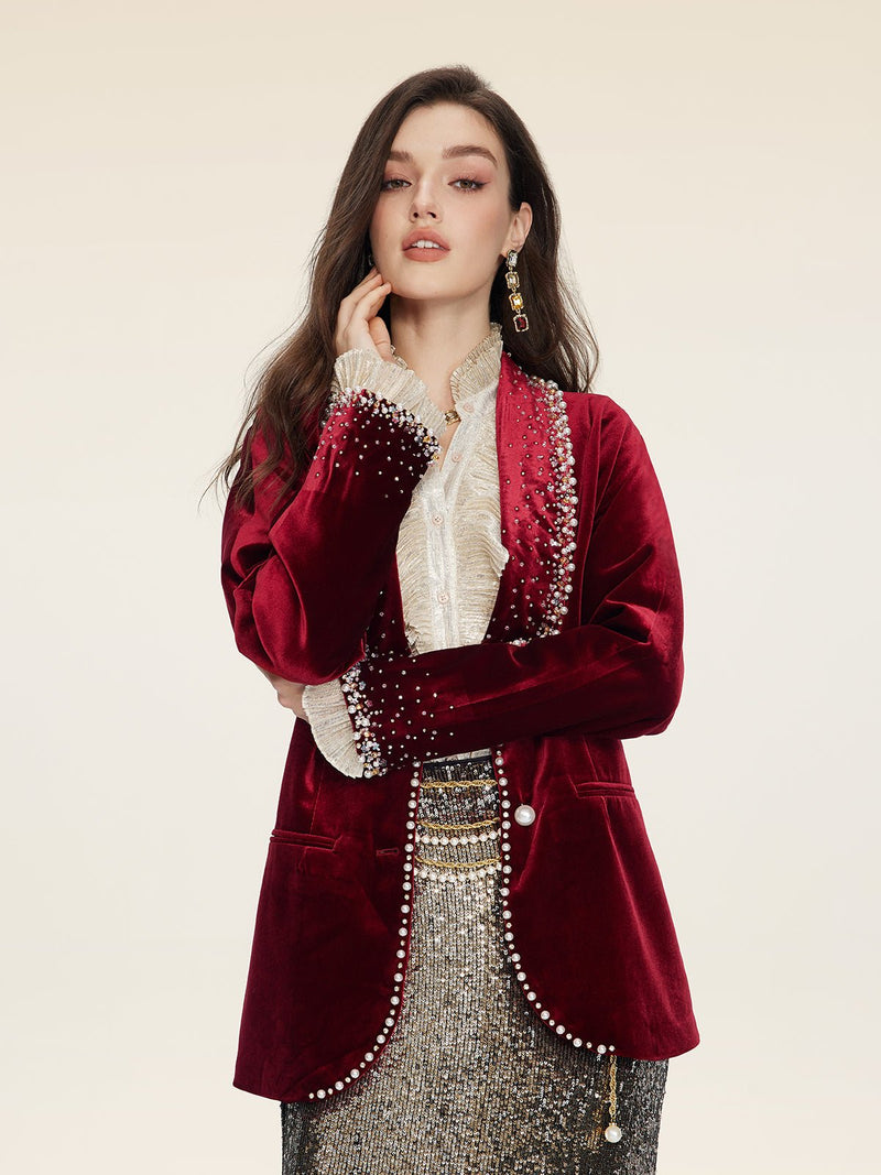 A woman wears the Social Queen Pearl & Crystal Velvet Blazer, adorned with hand-applied pearls, over a ruffled blouse and sequined skirt, evoking vintage glamour against a plain background.