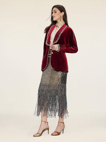 A woman stands in profile against a plain background, wearing Social Queen’s Pearl & Crystal Velvet Blazer styled over a metallic fringed skirt and gold heeled sandals.