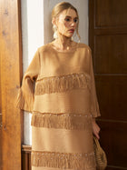 A woman stands indoors near wooden doors, holding a woven bag and wearing the Social Queen Pearl Detail Fringe Pleated Dress, featuring tan pleated chiffon, layered fringe, and pleated sleeves.