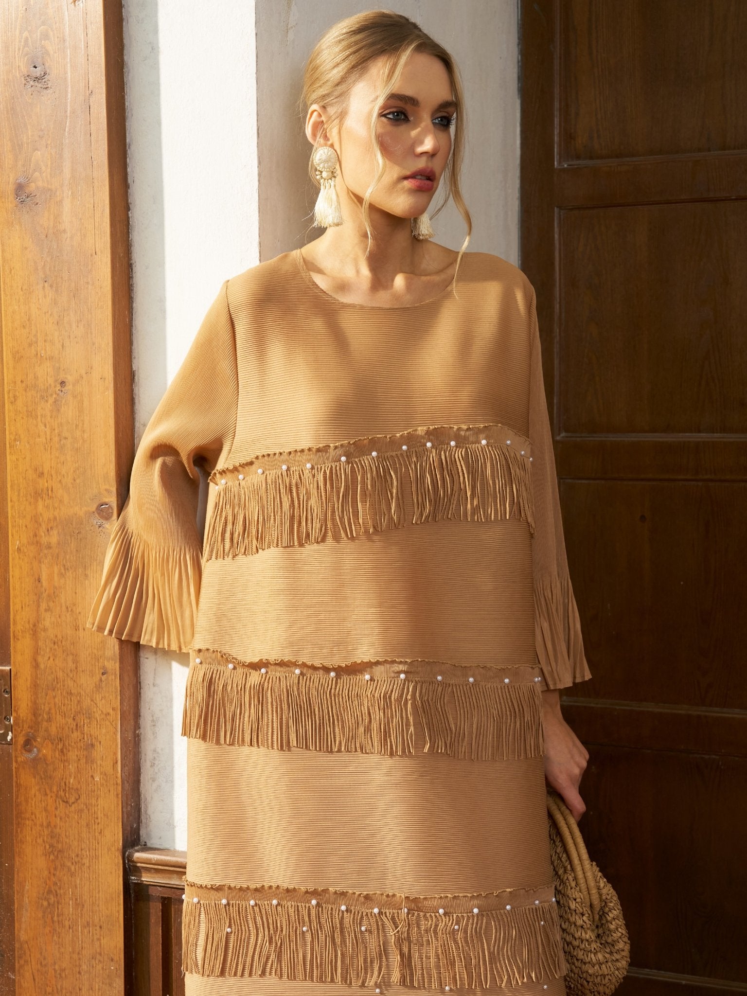 A woman stands indoors near wooden doors, holding a woven bag and wearing the Social Queen Pearl Detail Fringe Pleated Dress, featuring tan pleated chiffon, layered fringe, and pleated sleeves.