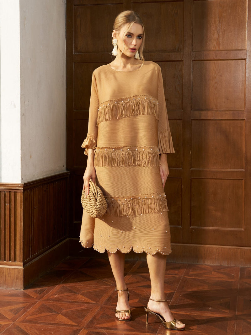 A woman wearing the Social Queen Pearl Detail Fringe Pleated Dress stands indoors against wooden paneling, holding a woven clutch.