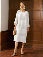 A woman wearing the Social Queen Pearl Detail Fringe Pleated Dress, featuring pleats and layered fringe, paired with a woven handbag, stands indoors against a wooden paneled wall.