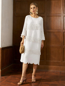 A woman wearing the Social Queen Pearl Detail Fringe Pleated Dress, featuring pleats and layered fringe, paired with a woven handbag, stands indoors against a wooden paneled wall.