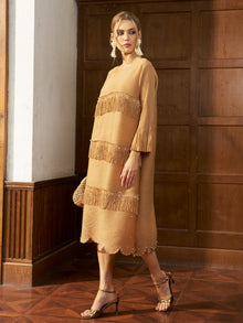 A woman stands indoors wearing the Social Queen Pearl Detail Fringe Pleated Dress, featuring tan long sleeves, layered fringe, and scalloped hems, styled with gold heels and a textured handbag.