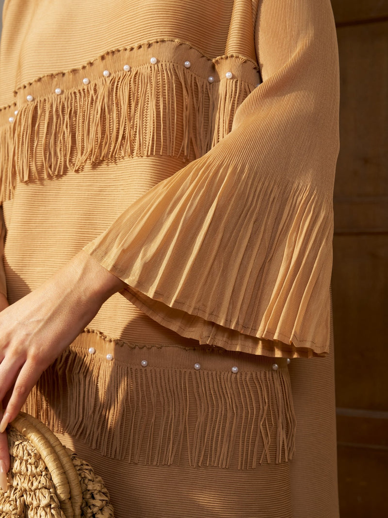 A close-up of a person wearing the Social Queen Pearl Detail Fringe Pleated Dress in tan, featuring pleated chiffon, layered fringe, faux pearl bell sleeves, and holding a woven straw handbag.