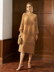 A woman stands indoors before a wooden paneled wall, wearing the Social Queen Pearl Detail Fringe Pleated Dress in tan. She holds a woven clutch and pairs the knee-length, pleated fringe dress with strappy heels.