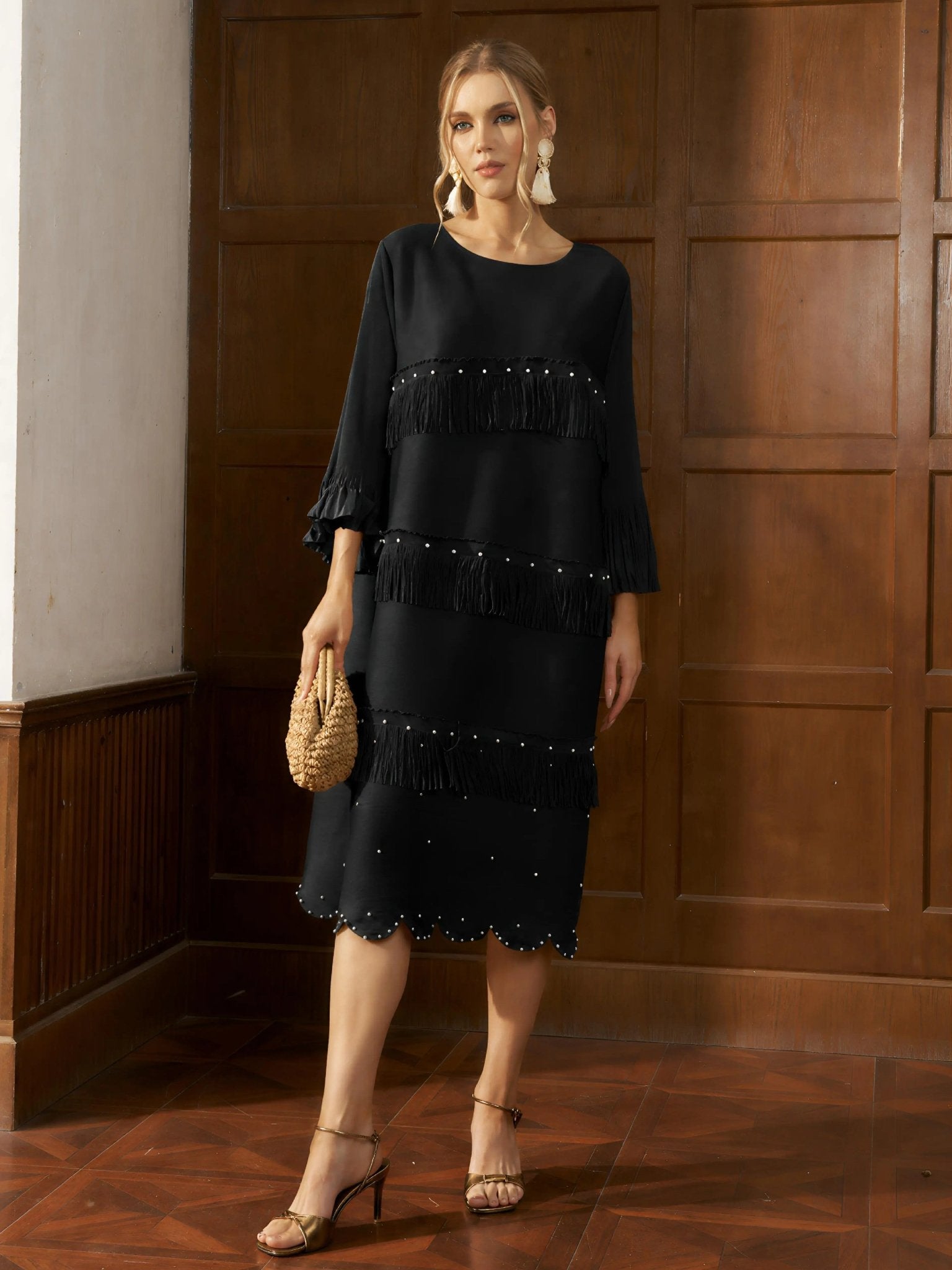A woman stands indoors wearing the Social Queen Pearl Detail Fringe Pleated Dress, a black, knee-length pleated chiffon style with layered fringe and bead accents, accessorized with a woven bag, strappy sandals, and statement earrings.
