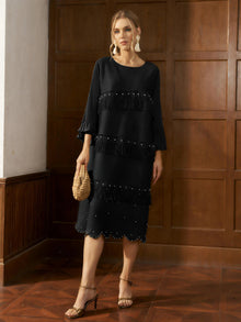 A woman stands indoors wearing the Social Queen Pearl Detail Fringe Pleated Dress, a black, knee-length pleated chiffon style with layered fringe and bead accents, accessorized with a woven bag, strappy sandals, and statement earrings.