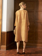 A woman stands indoors facing a wooden wall, wearing the Social Queen Pearl Detail Fringe Pleated Dress with layered fringe sleeves and holding a straw clutch.