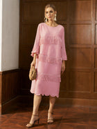 A woman wears the Social Queen Pearl Detail Fringe Pleated Dress—knee-length, pink, with pleats and fringe—holding a woven bag and standing indoors against a wooden panel wall.