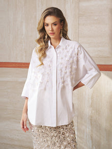 A woman wears the Pearl-Embellished Floral Blouse by Social Queen with a sequined skirt, creating an elegant look perfect for a wedding guest, as she stands against a marble wall.