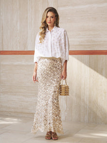 A woman indoors wears the Social Queen Pearl-Embellished Floral Blouse with a matching cape and sequin skirt, accessorized with a small handbag and gold sandals—the perfect wedding guest look.