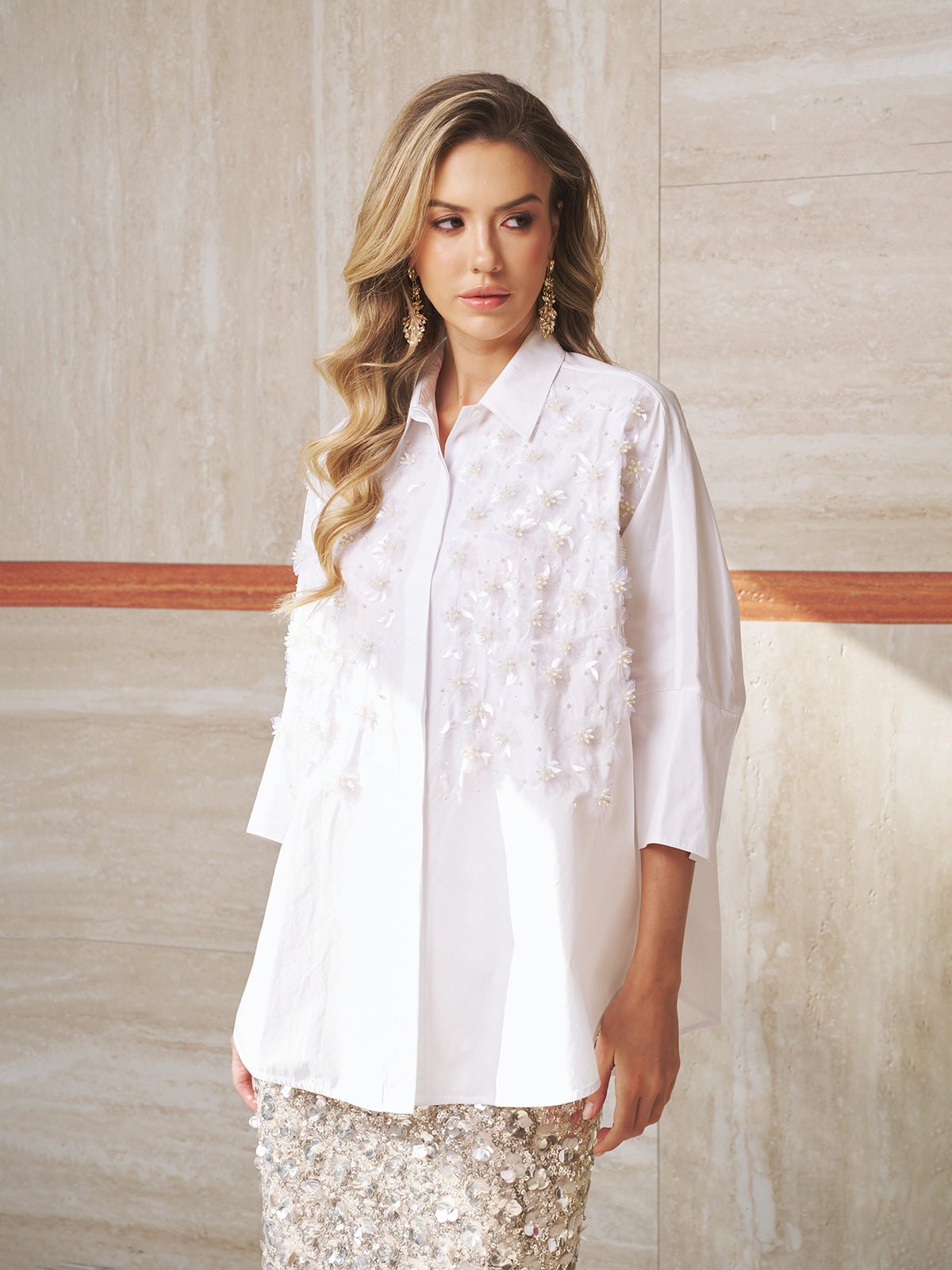 A woman with long wavy hair wears the Social Queen Pearl-Embellished Floral Blouse and stands against a beige stone wall.