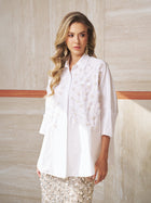 A woman with long wavy hair wears the Social Queen Pearl-Embellished Floral Blouse and stands against a beige stone wall.