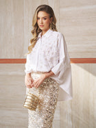 A woman wears the Social Queen Pearl-Embellished Floral Blouse with a sequined skirt and beige clutch, standing against a beige tiled wall.