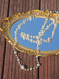 A gold-framed mirror rests on wood with the Social Queen Pearl Necklace with Crystal Accents draped across it, reflecting the sky.