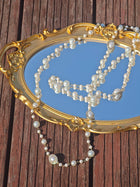 A gold-framed mirror rests on wood with the Social Queen Pearl Necklace with Crystal Accents draped across it, reflecting the sky.