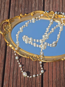 A gold-framed mirror rests on wood with the Social Queen Pearl Necklace with Crystal Accents draped across it, reflecting the sky.
