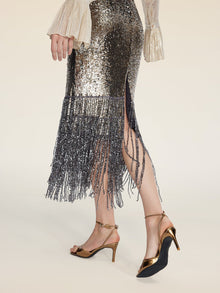 Wearing Social Queen's Pearl-Trim Sequin Fringe Skirt and gold high-heeled sandals, the person stands against a neutral background.