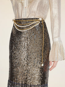 A person wears a shimmery blouse with flared cuffs and the Social Queen Pearl-Trim Sequin Fringe Skirt, accessorized with layered pearl chain waist belts.