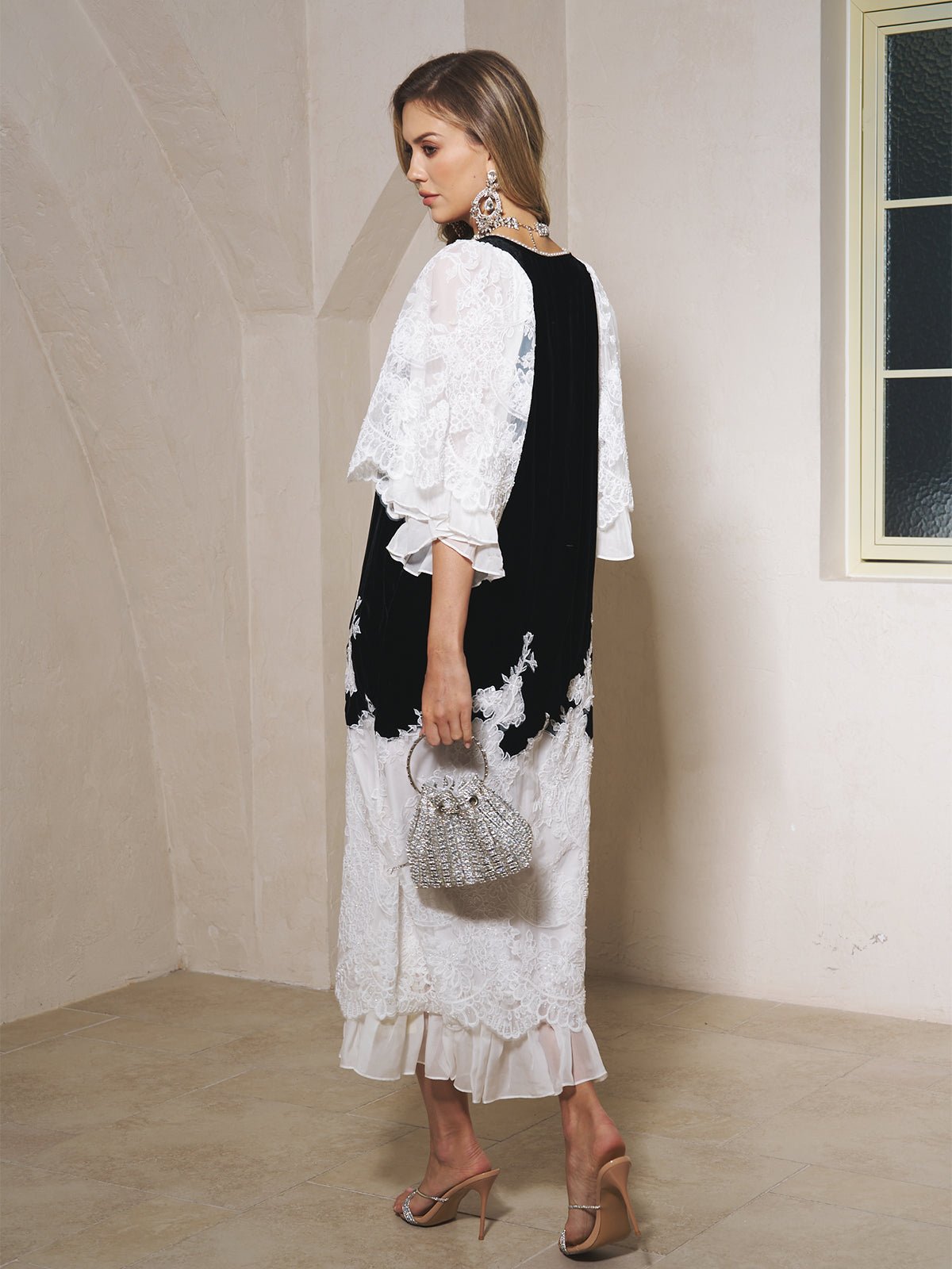 A woman stands indoors on a beige floor wearing the Social Queen Pearl-Trim Silk Velvet Dress, paired with heeled sandals, statement earrings, and a beaded handbag.