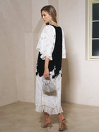 A woman stands indoors on a beige floor wearing the Social Queen Pearl-Trim Silk Velvet Dress, paired with heeled sandals, statement earrings, and a beaded handbag.