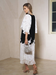 A woman stands indoors on a beige floor wearing the Social Queen Pearl-Trim Silk Velvet Dress, paired with heeled sandals, statement earrings, and a beaded handbag.