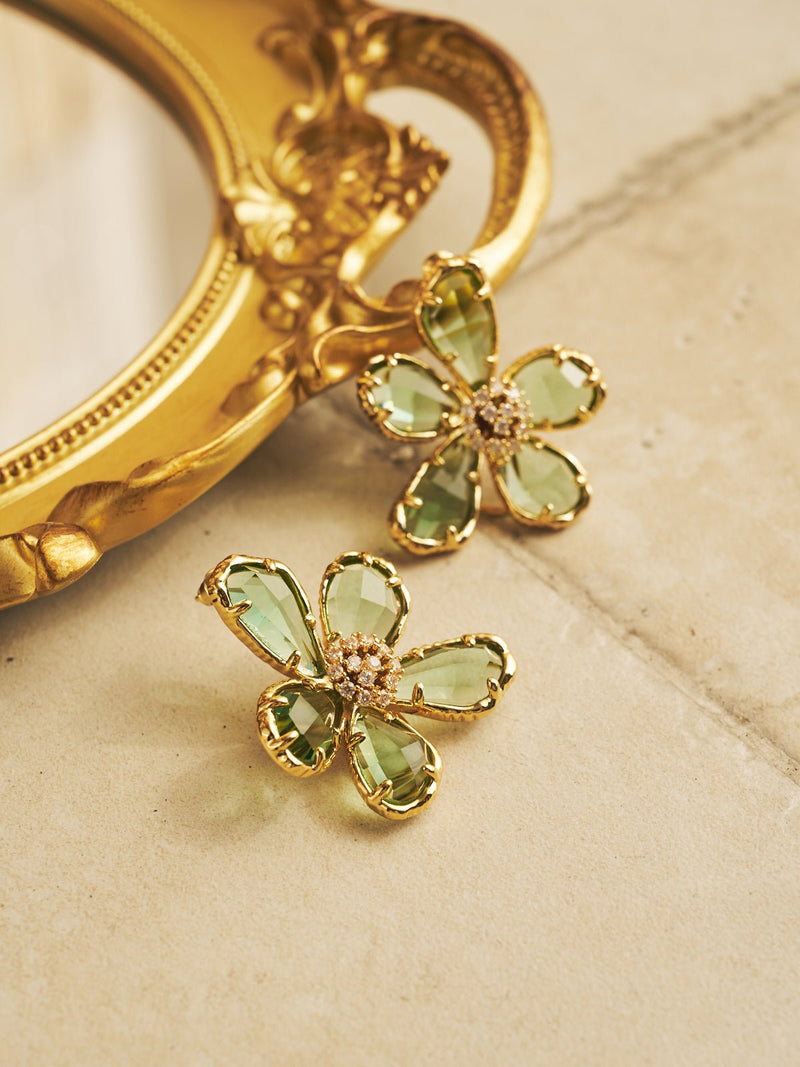 The Petal Earrings by Social Queen are gold-plated studs with a 3D flower design, green gemstone petals, and a rhinestone center, shown beside an ornate gold-framed mirror on a beige surface.