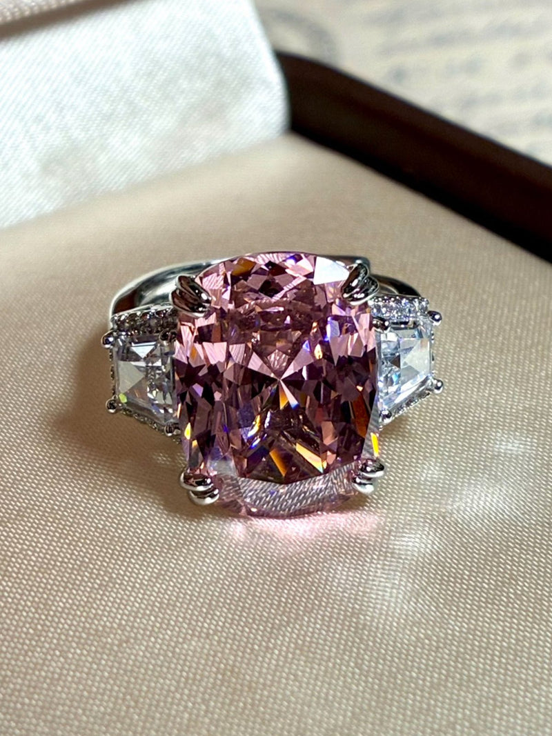 The Social Queen Pink Crystal Cushion Ring features a large rectangular pink crystal and two clear triangular stones, set in white gold plating and displayed on a beige surface.
