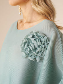 A woman models the Social Queen Pleated Floral Appliqué Set, featuring a textured light blue top with a bold 3D floral shoulder detail, paired with dangling pearl earrings.