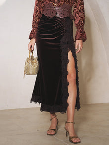A person wearing Social Queen's Pleated Lace-Panel Velvet Skirt, a patterned long-sleeve top, heeled sandals, and carrying a small beaded handbag.