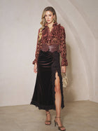 A woman stands indoors wearing the Social Queen Pleated Lace-Panel Velvet Skirt in black with a high slit and pleated waist, paired with a textured maroon blouse, gold heels, and carrying a small gold handbag.