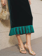 A person wears the Social Queen Pleated-Trim Velvet Stretch Skirt in black with an emerald pencil silhouette, gold heeled sandals, and a gold studded handbag while standing on a stone floor.