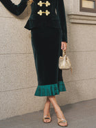 A person wearing a black velvet suit with gold buttons, the Social Queen Pleated-Trim Velvet Stretch Skirt featuring an emerald velvet accordion pleat trim, holding a small gold purse and wearing gold sandals.