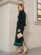 A woman wears Social Queen's Pleated-Trim Velvet Stretch Skirt in emerald, paired with gold heels and a small woven handbag, standing on a sidewalk near a stone building.