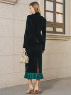 A woman stands against a stone wall in Social Queen's Pleated-Trim Velvet Stretch Skirt and matching emerald jacket, styled with gold heels and a small gold handbag.