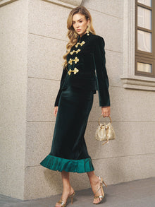 A woman in Social Queen’s Pleated-Trim Velvet Stretch Skirt, featuring gold buttons and accordion pleat trim, pairs it with a small gold handbag and gold heels as she poses by a stone wall and window.
