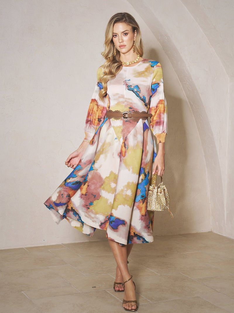 Indoors, a woman wears the Social Queen Pleated Wave Hem Art Print Dress with balloon sleeves and a belt, pairing it with heels and a textured gold handbag.