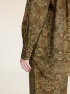A person models the Social Queen Printed Linen Blouse, featuring long sleeves and an intricate olive green pattern, shown from shoulder to mid-thigh with their hand by their side.