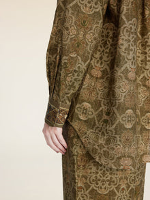 A person models the Social Queen Printed Linen Blouse, featuring long sleeves and an intricate olive green pattern, shown from shoulder to mid-thigh with their hand by their side.