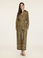 A woman stands facing forward wearing Social Queen’s Printed Linen Straight-Leg Pants in olive green with a patterned design, paired with earrings and heeled sandals.