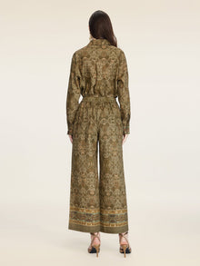 A person stands facing away, wearing Social Queen's Printed Linen Straight-Leg Pants in olive green with gold detailing, paired with a matching patterned long-sleeve top and open-toe heels.